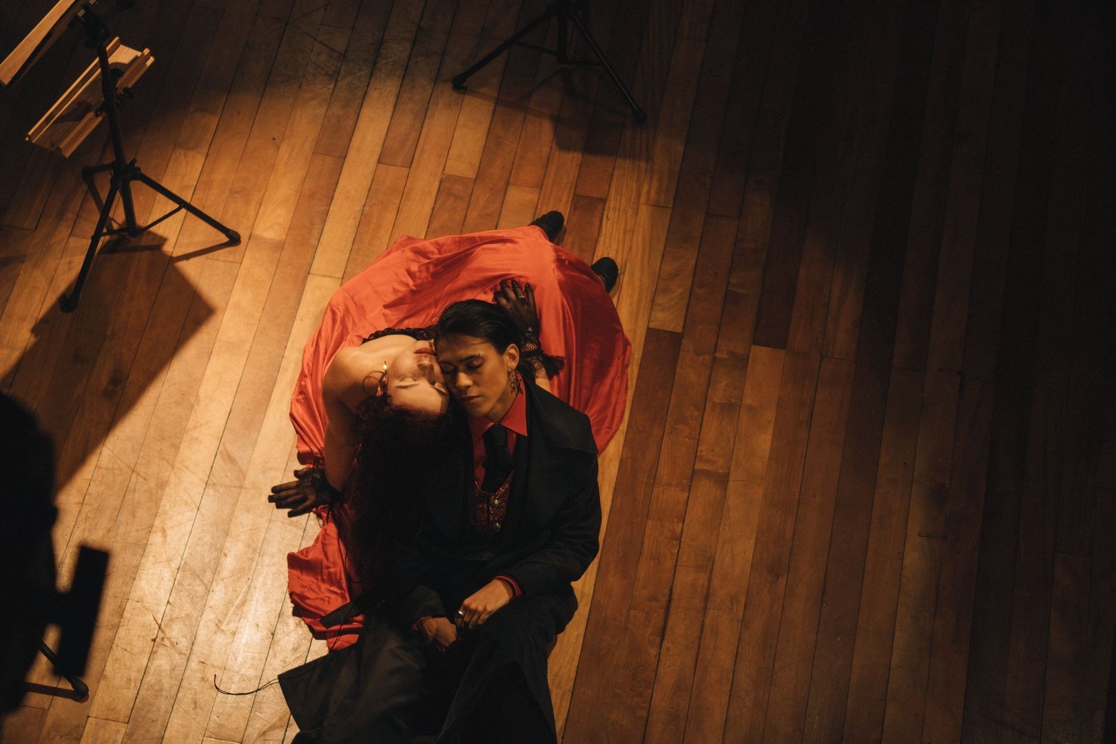 Two performers embrace on a warmly lit black box theater floor