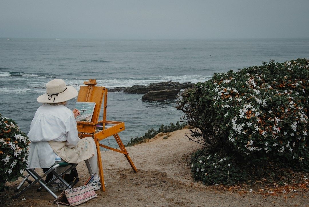 Artist painting at an easel beside a rocky, flower-lined coastline
