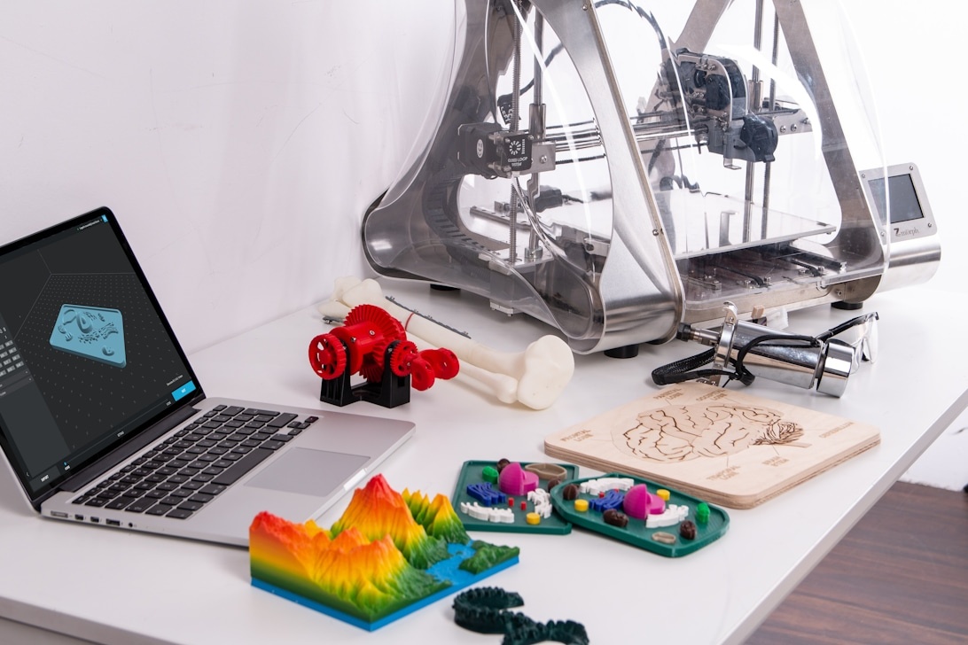 A comprehensive digital fabrication workspace featuring a 3D printer, laptop displaying CAD software, colorful 3D printed objects, and various tools representing modern digital art and design education.